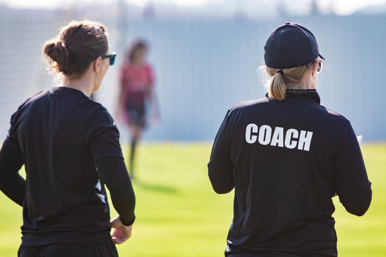Back view of female sport coach and her assistant in black COACH shirt at an outdoor sport field, watching their girl football team